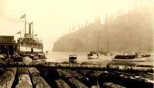 Terminal Steam Navigation Co. steamer “Baramba” at Bowen Island Wharf with log boom in foreground and boats at anchor to the right. ca.1910